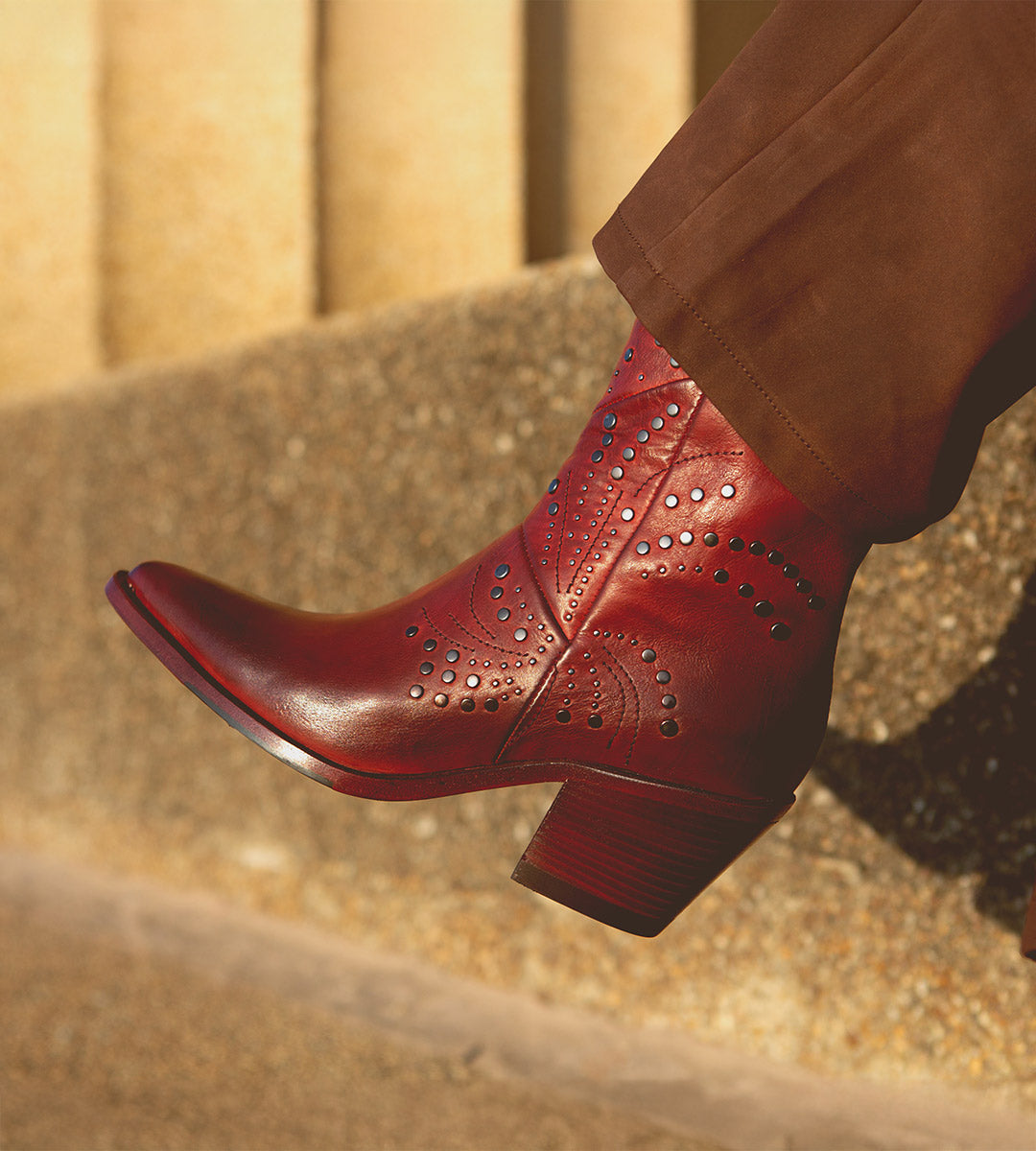A person wearing brown pants and red Sposa ankle booties by Bed Stu, featuring decorative patterns, pointed toes, and studs, stands on a textured stone surface.