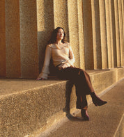 A woman wearing a beige sweater and dark pants, complemented by Bed Stu Sposa Italian leather booties with pointed toes, sits against a large stone structure, bathed in warm sunlight.