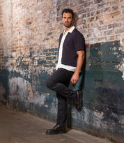 A man with short dark hair and a beard leans against a weathered brick wall, wearing a white shirt, dark short-sleeve jacket, black jeans, and Bed Stu Tempt hybrid Chelsea boots.