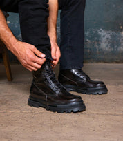 Wearing black pants, a person ties the laces of Bed Stu Tempt boots—stylish hybrids mixing hiking-boot ruggedness with modern flair—while seated on a wooden chair atop a concrete floor.