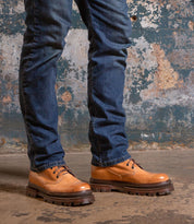 A person in blue jeans stands on a concrete floor by a weathered wall, wearing Bed Stu’s Tempt—a stylish hybrid shoe that blends the classic Chelsea boot look with rugged, thick soles.