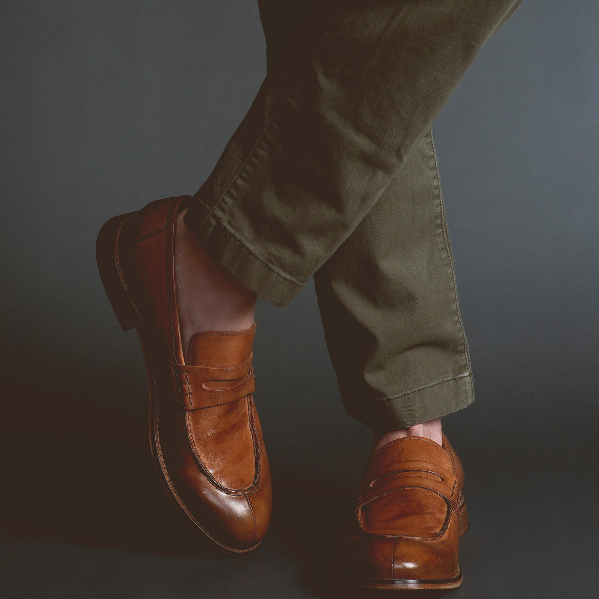 A person wearing handcrafted brown Time loafers by Bed Stu and olive green pants stands against a dark background, exuding sophistication.