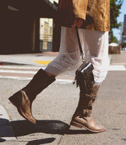 A person walking on the street is wearing a brown jacket, a sheer white dress with white ruffle trim, and Bed Stu's Turn brown leather boots made with Goodyear welted construction, while holding a small black handbag.