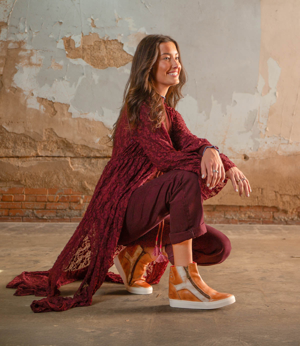 A woman wearing Bed Stu's Twist tan high-top sneakers with hidden wedge crouches against a worn, textured wall, pairing them with a long maroon top and burgundy pants.