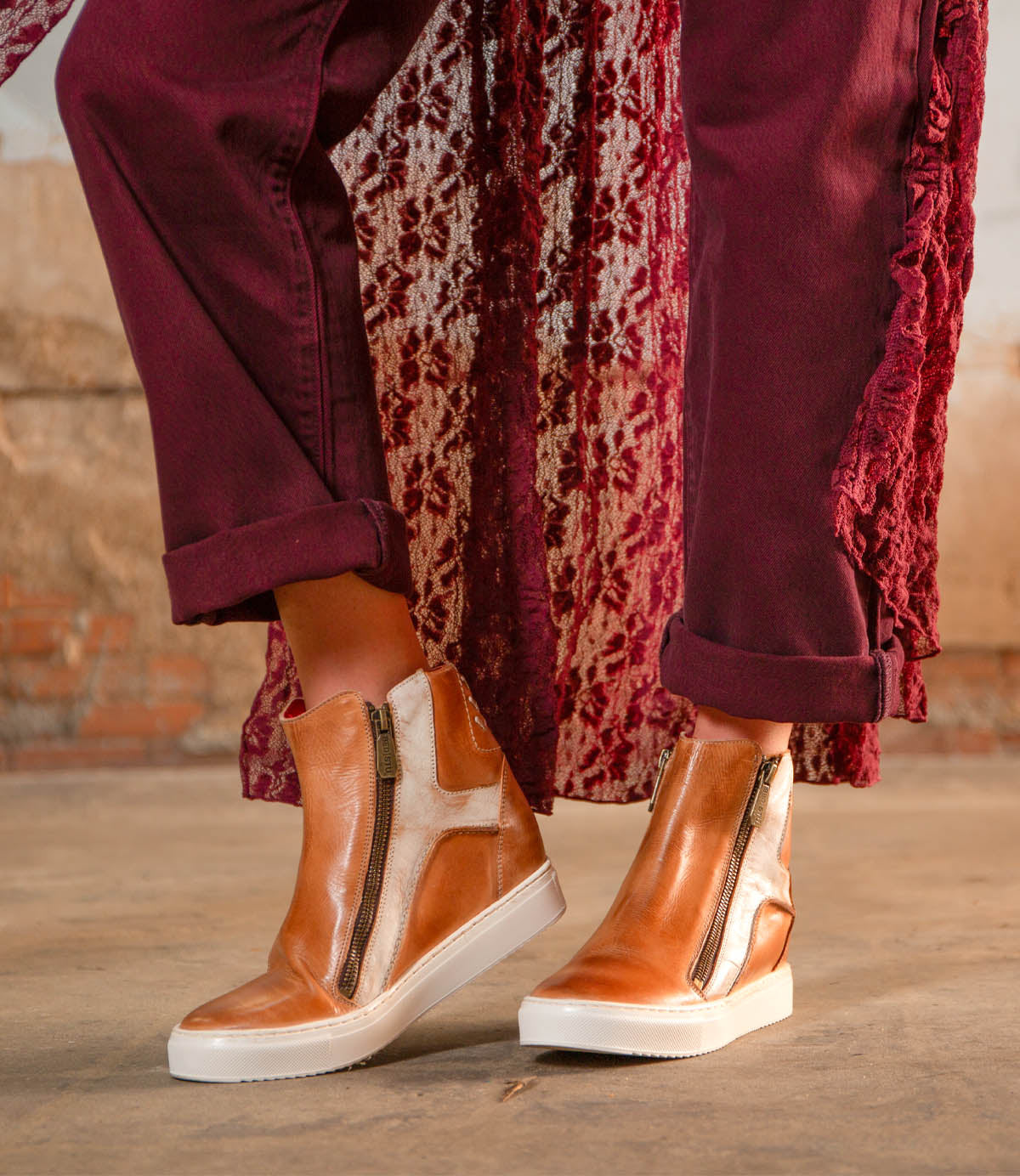 A person stands on a concrete floor wearing Bed Stu's Twist tan high-top sneakers with leather panels and side zippers, paired with burgundy pants and a textured burgundy outer layer.