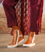 A person stands on a concrete floor wearing Bed Stu's Twist tan high-top sneakers with leather panels and side zippers, paired with burgundy pants and a textured burgundy outer layer.