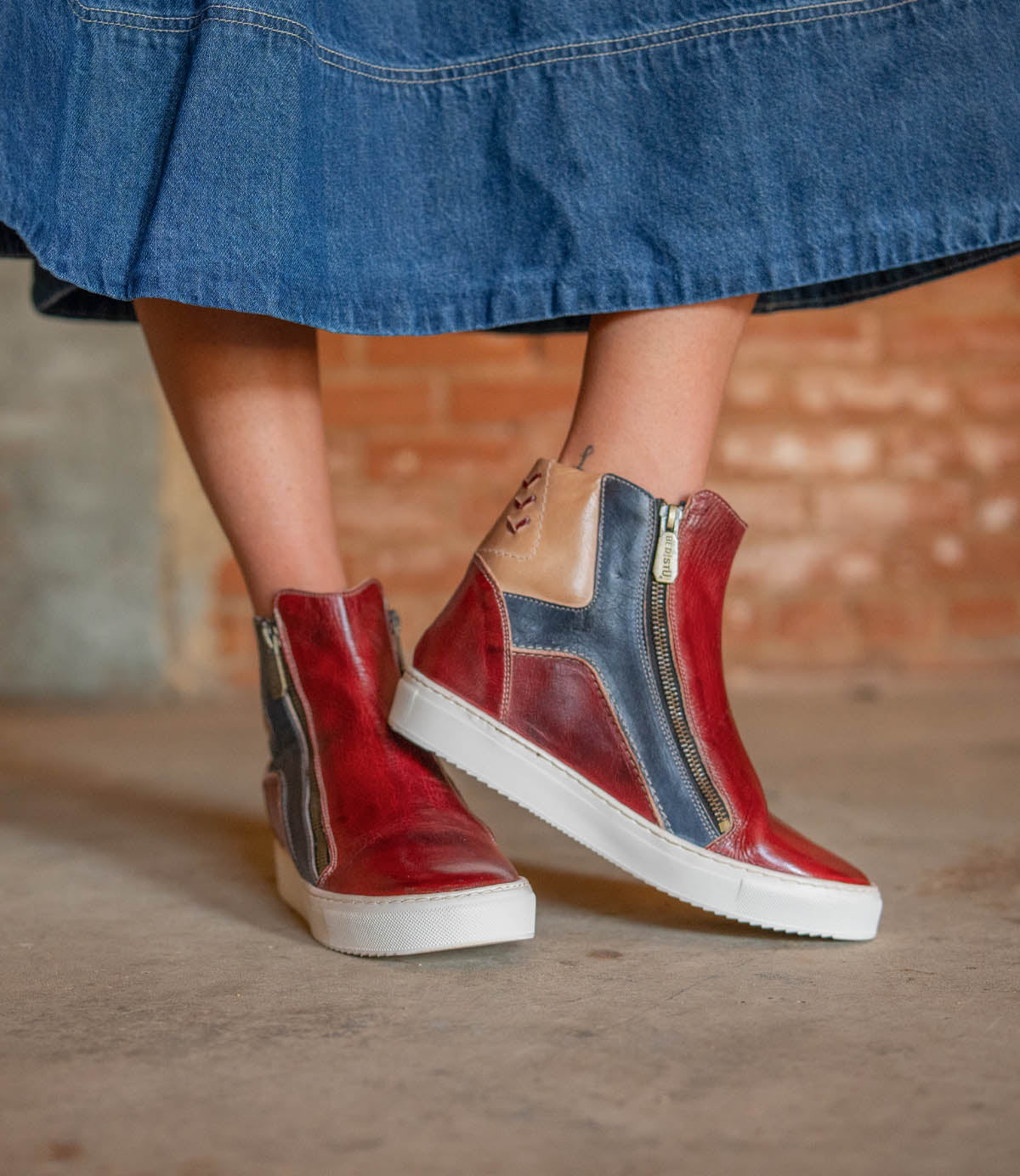 A person wears the Bed Stu Twist high-top sneakers featuring leather panels and side zippers, paired with a blue denim skirt, standing on a concrete floor before a brick wall.