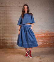 A woman stands against a brick wall wearing a denim outfit with red Twist high-top sneakers by Bed Stu, featuring sleek leather panels.