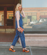 Woman walking in Bed Stu denim overalls and environmentally-conscious Bed Stu Vanquish mules, holding a small purse, with a street background.