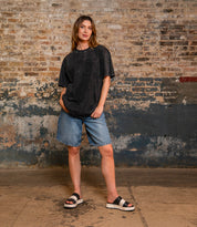 A woman stands indoors by a brick wall, wearing a loose dark tee, denim shorts, and Bed Stu’s Vext platform slide sandals with a leather-wrapped sole.