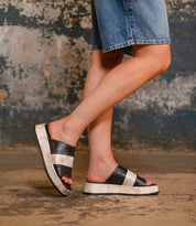 A person wears Bed Stu's Vext black and white leather-wrapped platform slide sandals with denim shorts, standing on a textured concrete floor against a weathered wall.