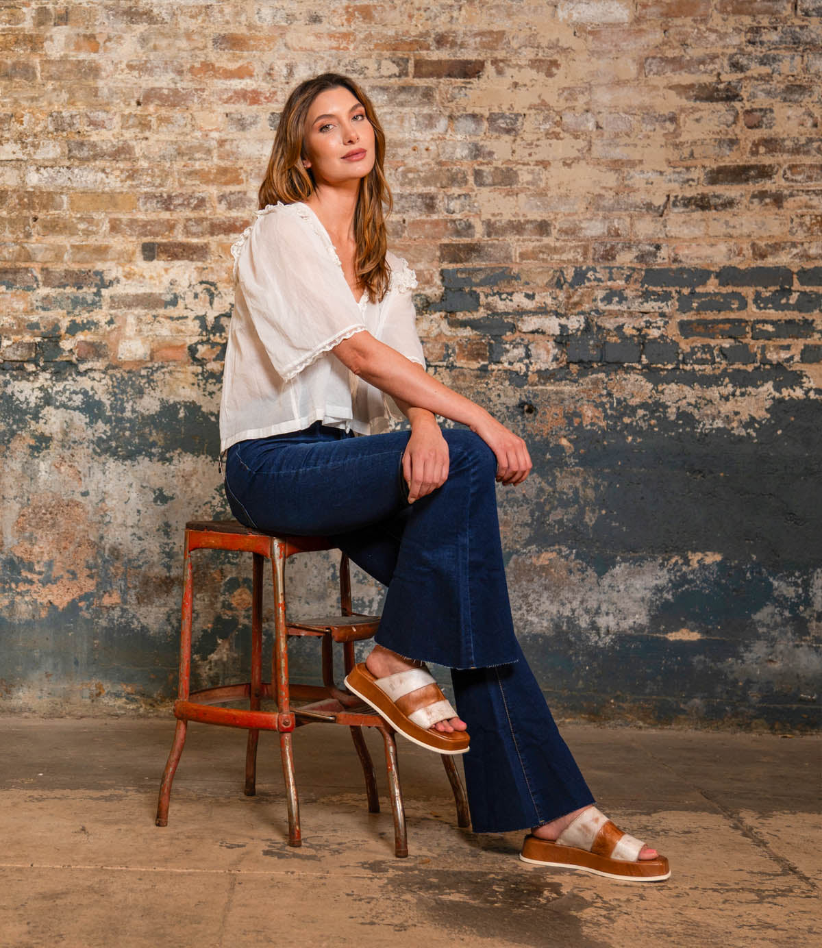 A woman sits on a rustic stool against a weathered brick wall, wearing a white blouse, blue flared jeans, and the Bed Stu Vext platform slide sandals with a leather-wrapped sole.
