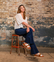 A woman sits on a rustic stool against a weathered brick wall, wearing a white blouse, blue flared jeans, and the Bed Stu Vext platform slide sandals with a leather-wrapped sole.