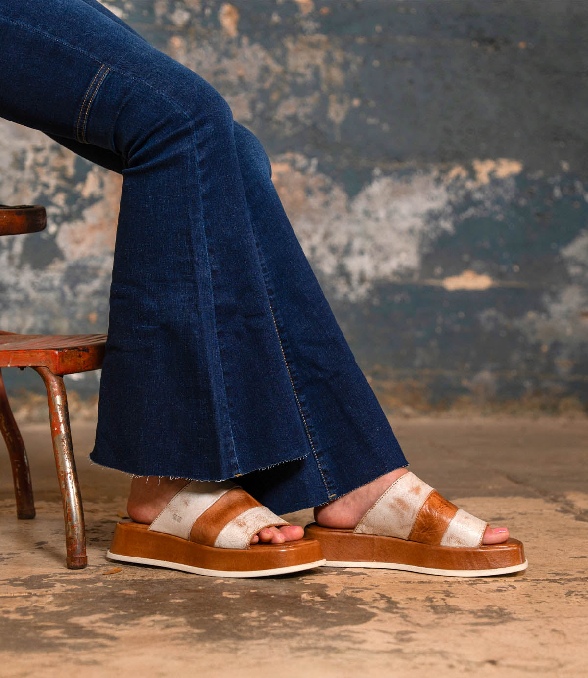 A person wearing flared blue jeans and Bed Stu Vext brown and white platform slide sandals with a leather-wrapped sole sits on a rustic metal chair against a textured wall.
