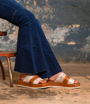 A person wearing flared blue jeans and Bed Stu Vext brown and white platform slide sandals with a leather-wrapped sole sits on a rustic metal chair against a textured wall.