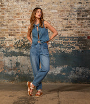 A woman in the Bed Stu Vice denim sleeveless top and jeans poses by a textured brick wall, looking to the side with one hand on her hip and chic strappy sandals on.