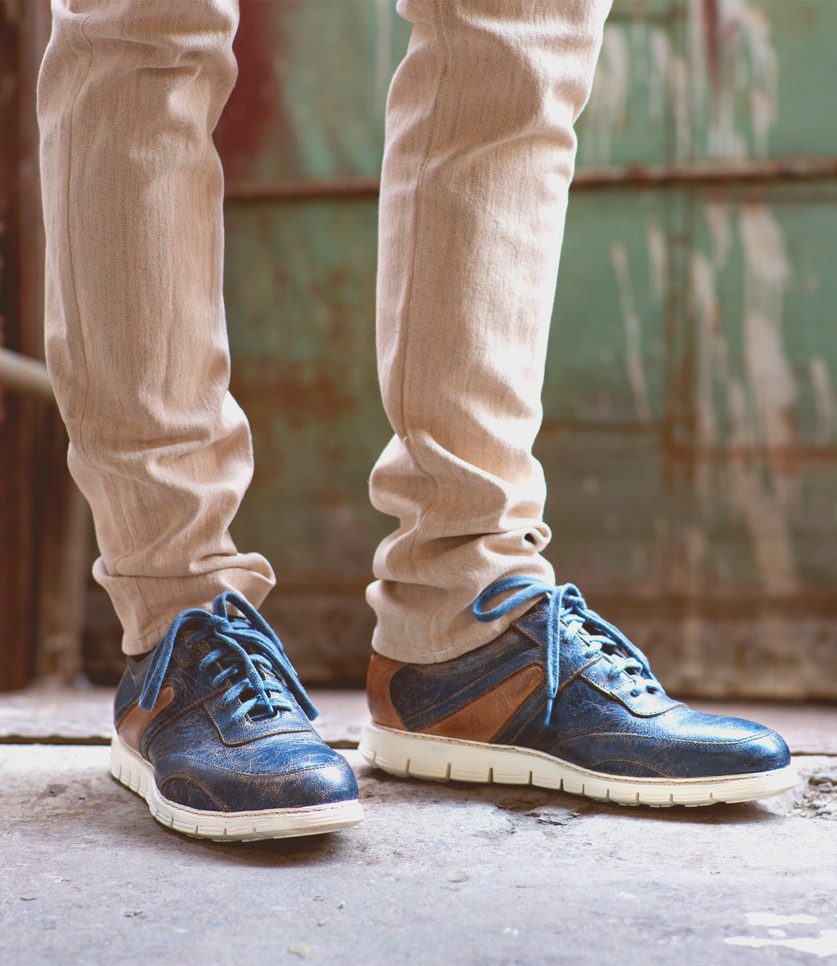 A person standing in an urban setting, wearing distressed Bed Stu lace-up sneakers and beige pants, with a rusted green door in the background.