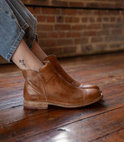 A person wears Bed Stu Yurisa light brown leather ankle boots with rolled-up jeans, sitting on a wooden floor by a brick wall. The everyday boot features a red leather lining and reveals a small flower tattoo on the ankle.