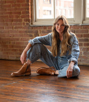 A woman in a denim jumpsuit sits on a wooden floor by a brick wall and window, wearing Bed Stu Yurisa brown ankle boots with a Vibram outsole and red leather lining, one leg bent in relaxed style.
