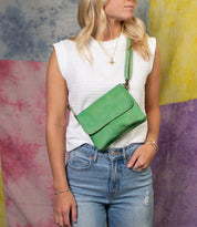 A person in a white sleeveless top and jeans poses with the Bed Stu Ziggy green leather crossbody bag, enjoying hands-free convenience against a colorful, textured background.