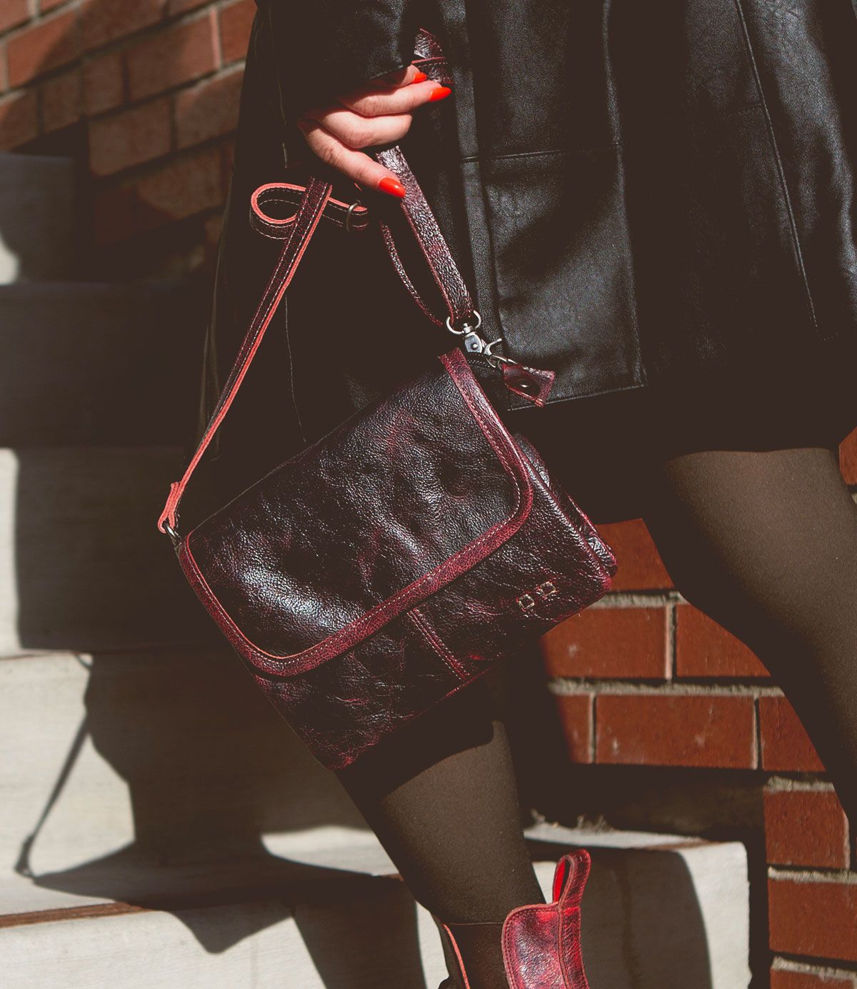 Person wearing dark clothing holding a Bed Stu Ziggy dark red leather crossbody handbag with a matching strap while standing on stairs near a brick wall.