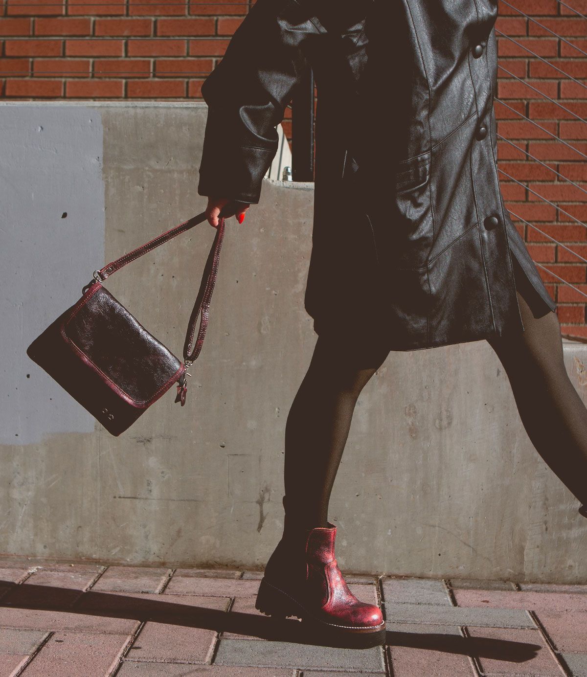 A person in a black leather coat, black leggings, and a pair of Bed Stu's Ziggy red boots strides along a sidewalk, holding a trendy belt bag with a brick wall in the background.