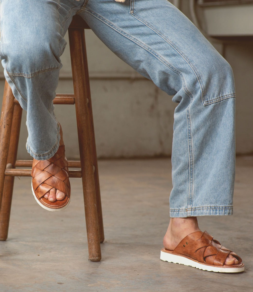 A person wearing Bed Stu Abraham Light Black Lux Sandals with a woven upper and blue jeans seated on a wooden stool, with one foot resting on the stool's lower rung.