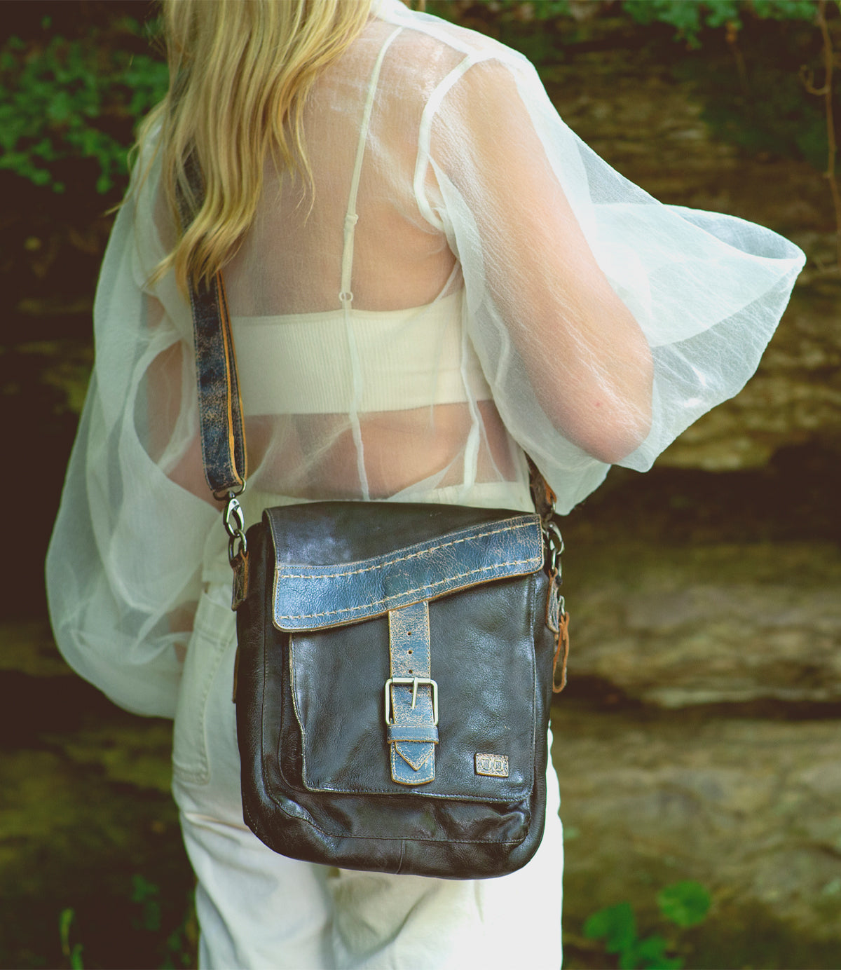 A woman with long blonde hair seen from behind wearing a sheer white shawl and white pants, carrying a Bed Stu Ainhoa LTC dark leather crossbody handbag.