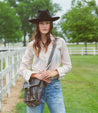 A woman in a black hat, polka dot blouse, and denim jeans stands by a white fence with a Bed Stu Ainhoa LTC crossbody handbag, in a rural setting.