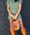 A woman in a green jumpsuit and brown boots holding a Bed Stu Ainhoa LTC leather crossbody handbag, standing against a textured dark brick wall. Only her torso and legs are visible.