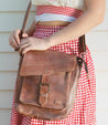 A woman wearing a red and white checkered skirt, holding a Bed Stu Ainhoa LTC brown leather crossbody handbag against a wooden background.