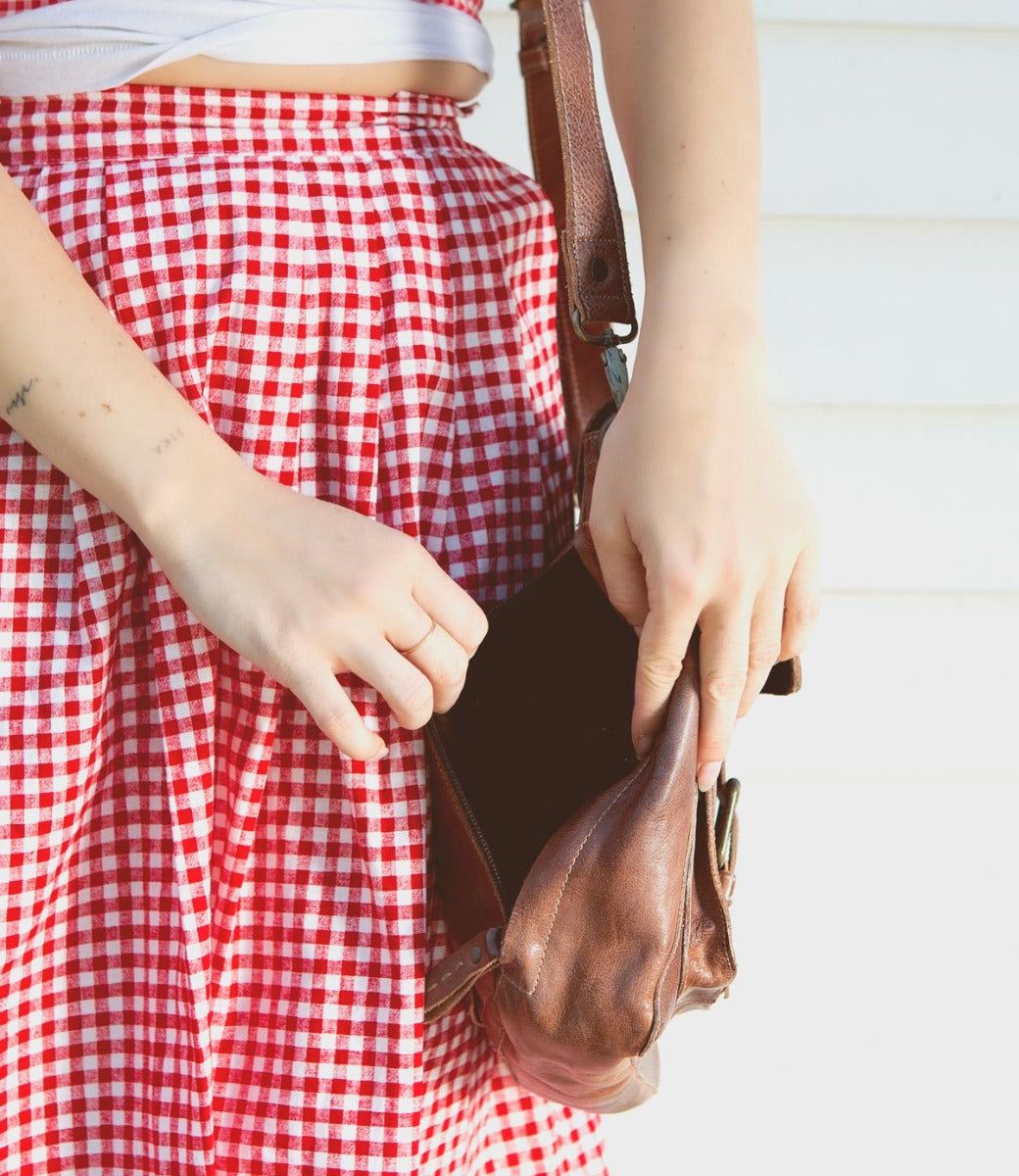 Woman in a red and white checkered dress taking a book out of a Bed Stu Ainhoa LTC brown leather crossbody handbag.