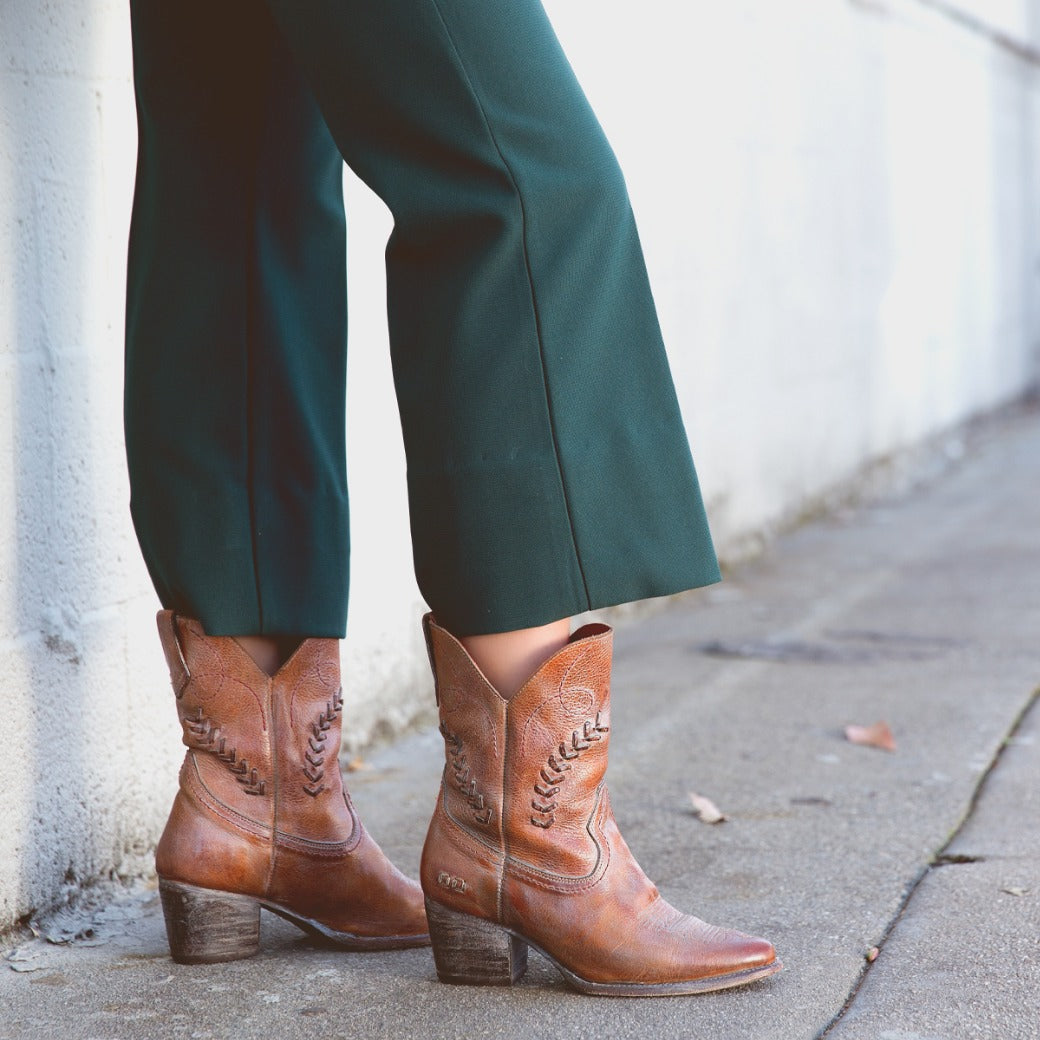A woman wearing green pants and brown Bed Stu cowboy boots.