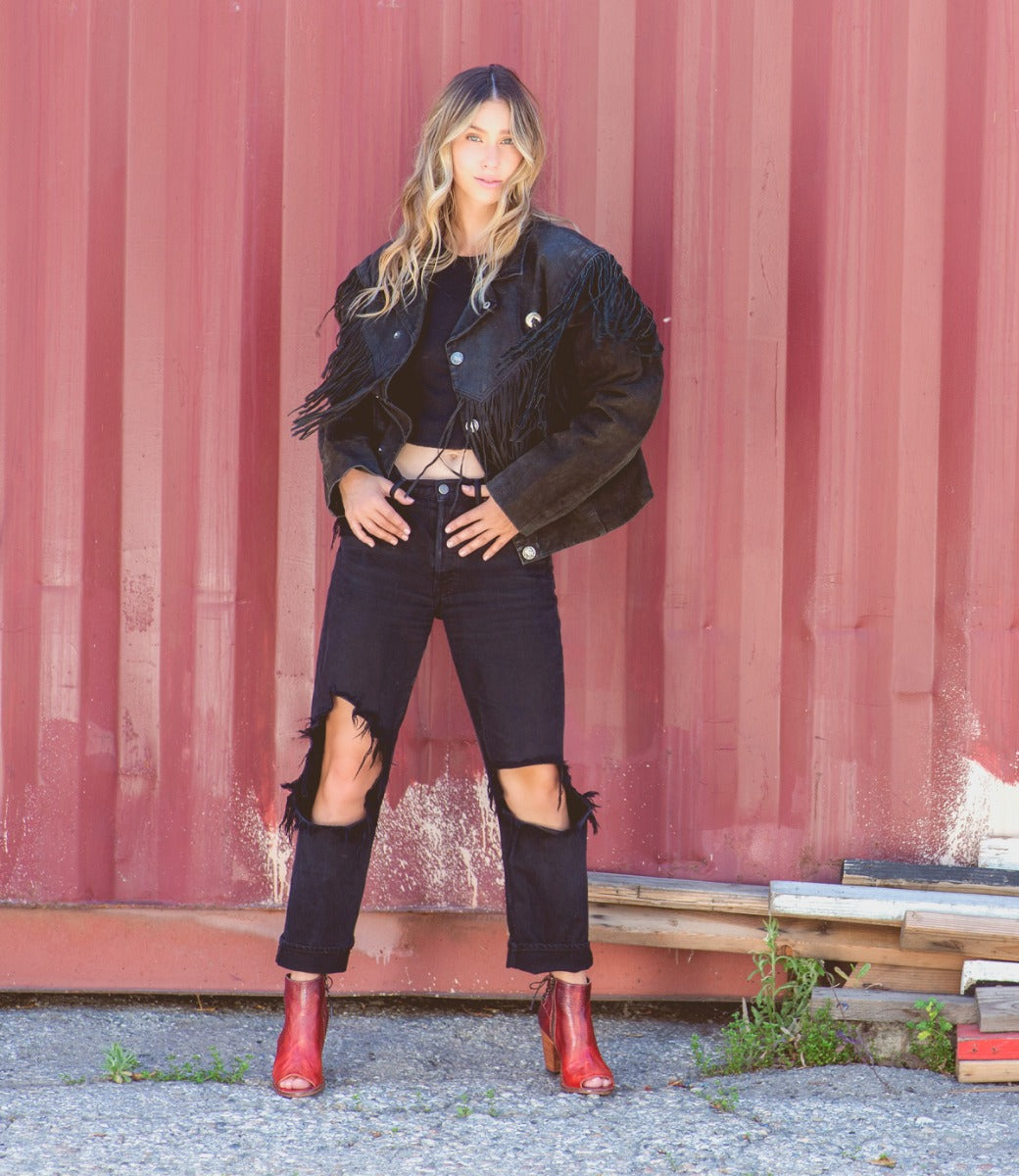 A woman in black jeans and red boots, the Angelique by Bed Stu, is posing in front of a red wall.