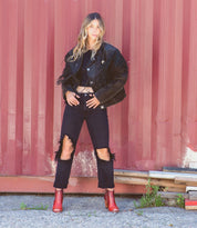 A woman in black jeans and red boots, the Angelique by Bed Stu, is posing in front of a red wall.
