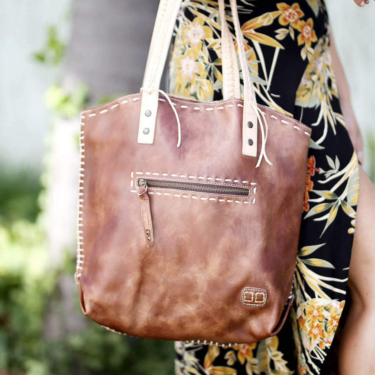 A close-up of a brown Bed Stu Barra handmade leather tote bag with contrast stitch detail and a zipper pocket, held by a person wearing a floral dress.