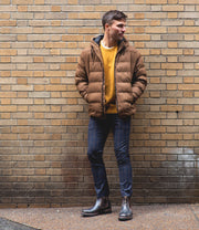 Man standing against a brick wall, wearing a brown puffer jacket, yellow sweater, plaid pants, and Bed Stu Brady Trek Black Lux Boot.