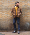 Man standing against a brick wall, wearing a brown puffer jacket, yellow sweater, plaid pants, and Bed Stu Brady Trek Black Lux Boot.