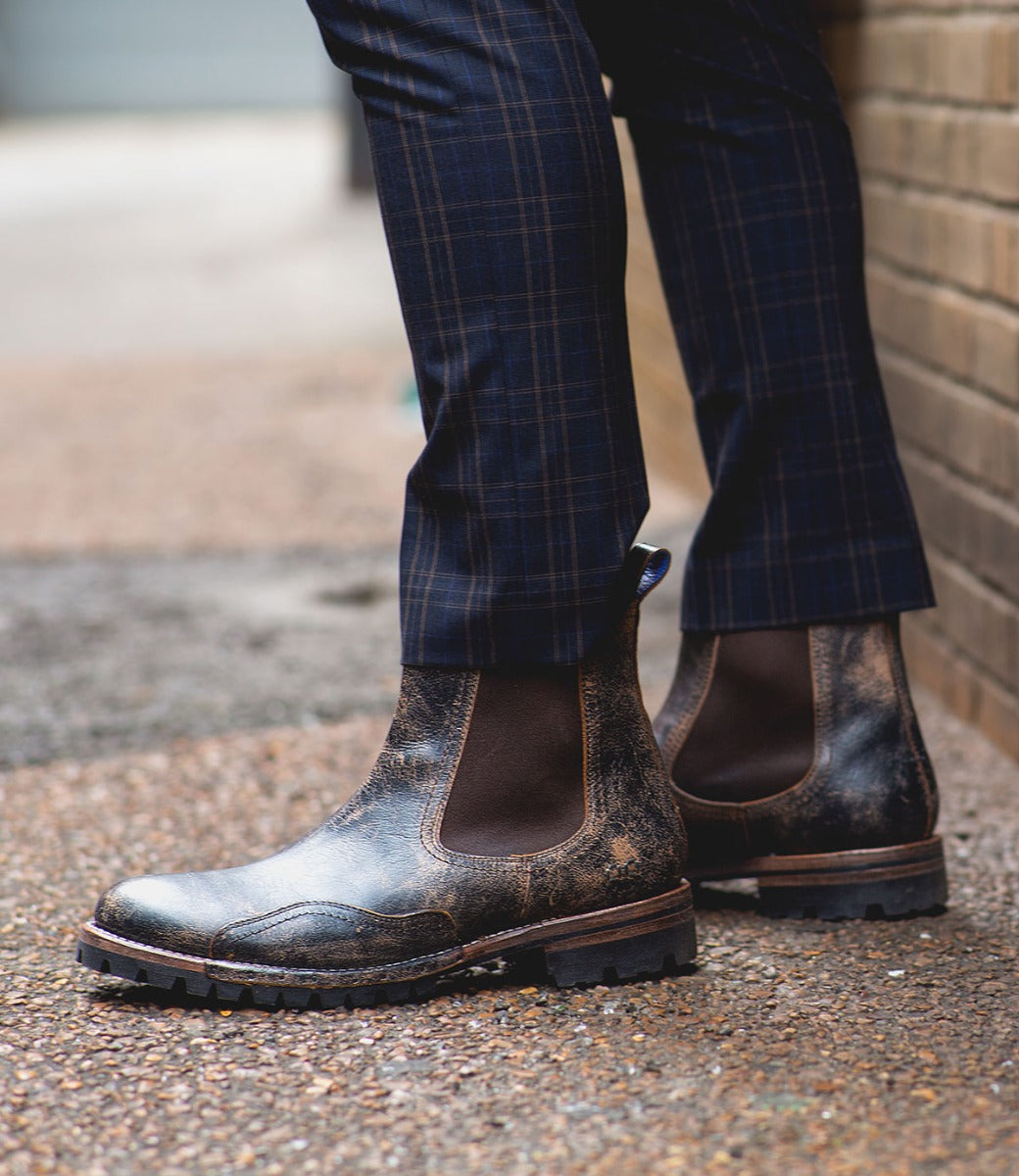 A person wearing Bed Stu Brady Trek boots with a Vibram outsole and plaid trousers standing on a brick-lined sidewalk.
