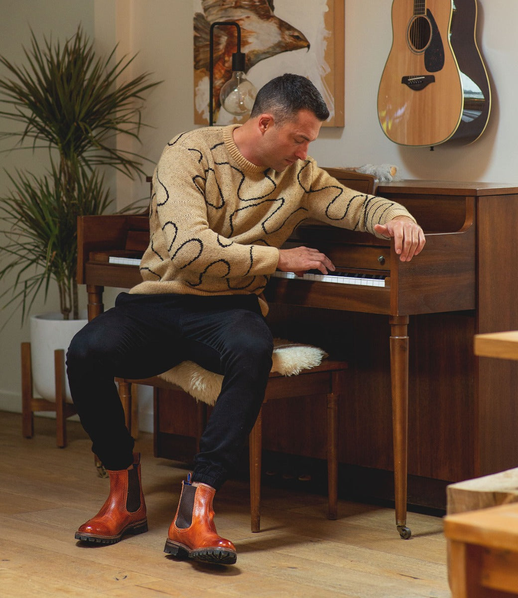 A man in a patterned sweater and Bed Stu Chelsea boots sitting at a piano in a cozy room with a guitar hanging on the wall and plants in the background.