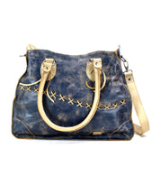 The Bruna tote bag from Bed Stu is a blue leather accessory with beige handles and intricate cross-stitch patterns. Displaying artisanal craftsmanship, this leather bag features a detachable shoulder strap with silver hardware.