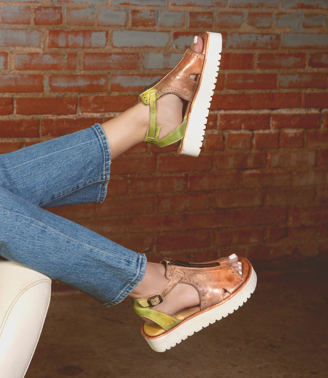Feet in Bed Stu's Clancy sandals, metallic copper and green with cushioned insoles and white platform soles, rest on a beige chair. Denim jeans against a brick wall complete the scene.