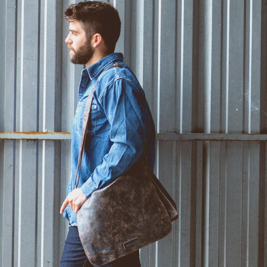 A man wearing a blue shirt and jeans is carrying a Bed Stu Hampton II messenger bag.
