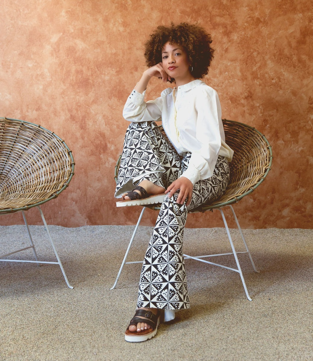 A woman sits on a chair wearing Bed Stu patterned pants and a white shirt.