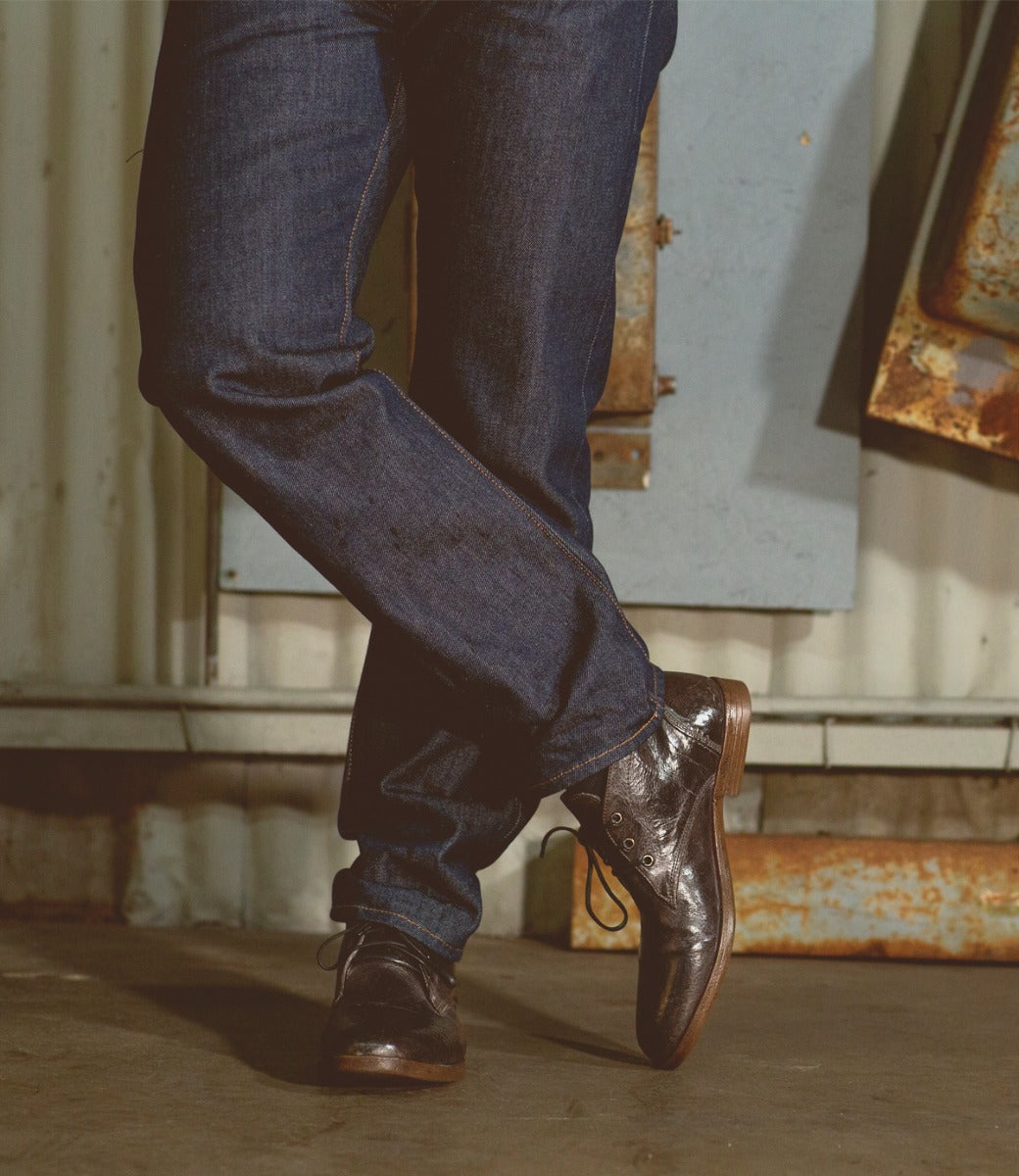 Close-up of a person's lower legs and feet, wearing dark jeans and brown leather Bed Stu Illiad chukka boots, standing in a dimly lit, industrial setting.