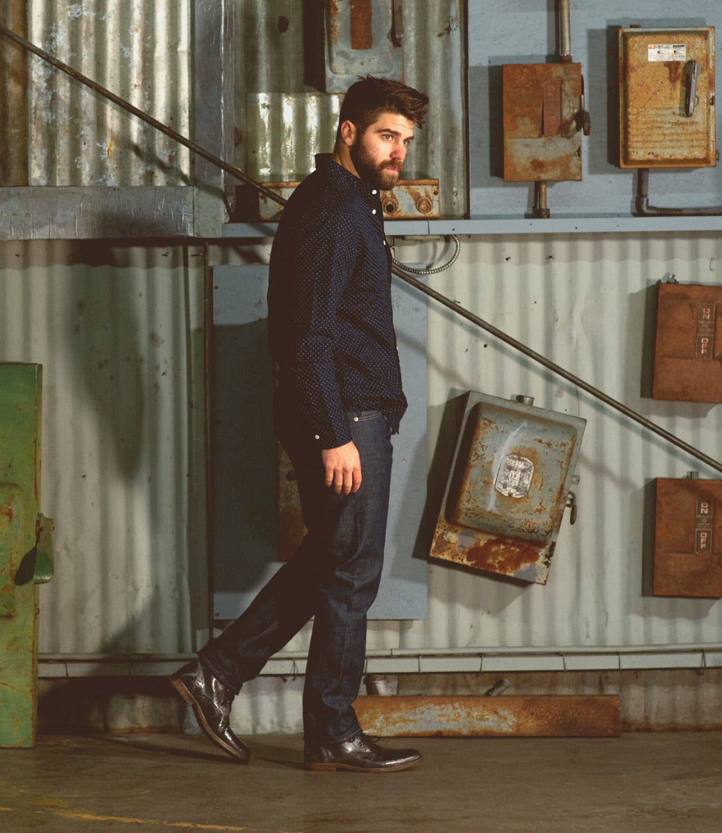 A man with a beard wearing jeans and a Bed Stu Illiad boot stands beside metal containers in a dimly-lit industrial setting.