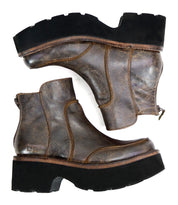 A pair of Bed Stu Infamy boots, made from brown leather with prominent stitching and a chunky outsole, showcasing a textured surface and equipped with side zippers, positioned side by side on a white background.