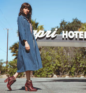 A woman in an Isla denim dress standing in front of a hotel sign. (Brand: Bed Stu)
