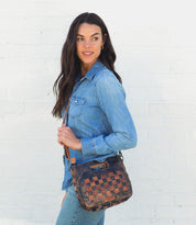 A woman wearing a denim shirt, jeans, and black and brown woven Keiki bag while standing next to a white wall.