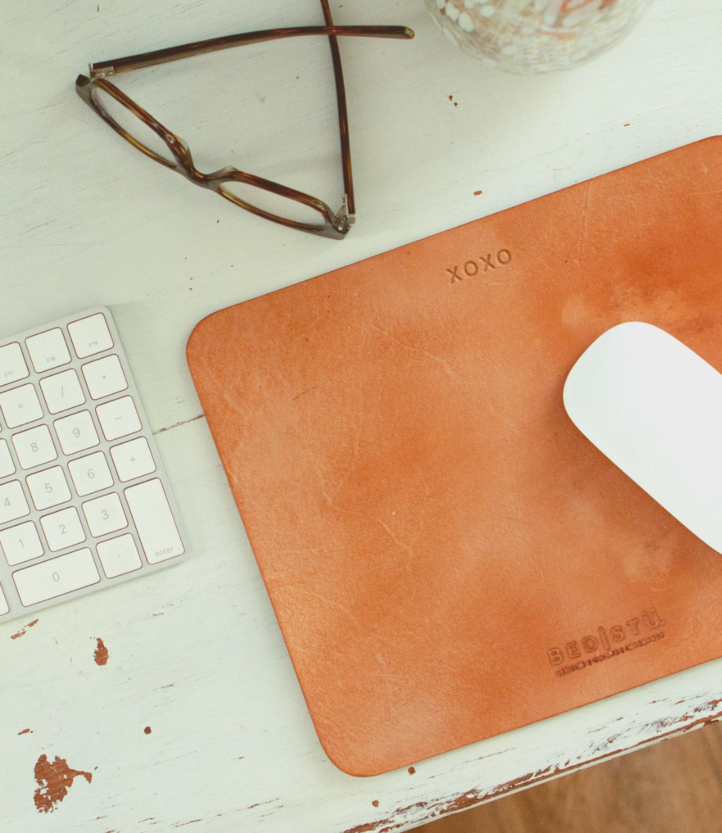 A beige leather Launcher Mouse Pad by Bed Stu with a suede underside, adorned with "XOXO" and "BED|STÜ" written on it, a white computer mouse, a keyboard, and a pair of glasses are neatly arranged on the curated workspace's white desk.